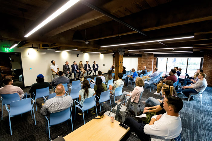 Shot from the back corner of a large classroom with approximately 20 people facing a panel of speakers at the front of the classroom