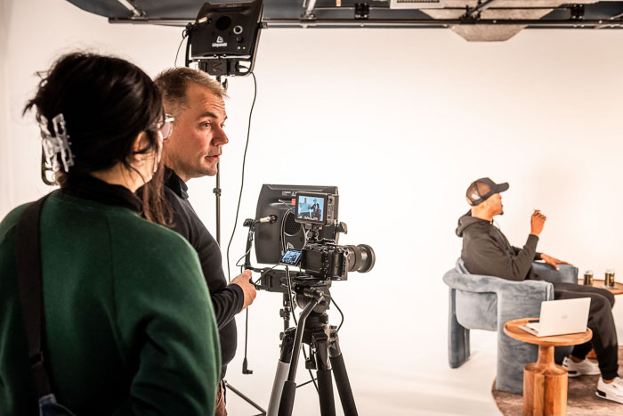 Two people stand in front of a camera in a white studio room.