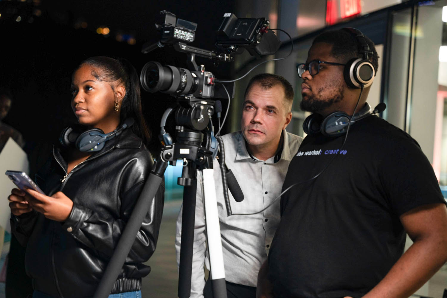 Three people stand around a video camera on a tripod. Two of them look at the camera while the third person, at the left of the frame, looks off to the distant holding a cell phone.