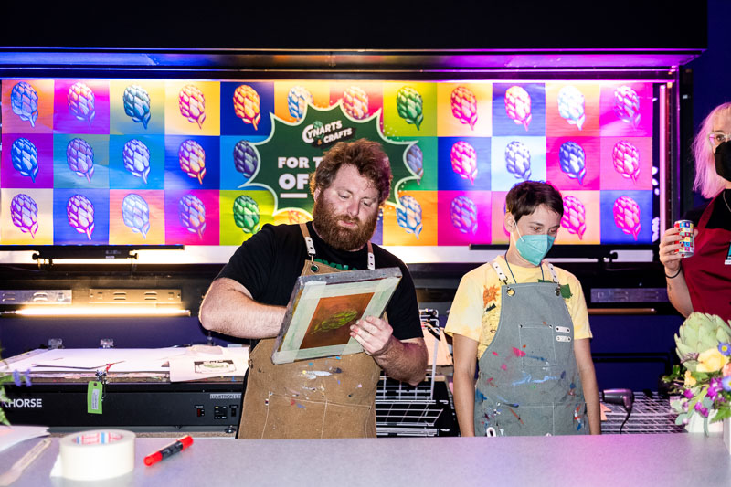 Two white people stand against a neon background featuring a repeating graphic of hops. The person in the center is holding a screenprinting screen.