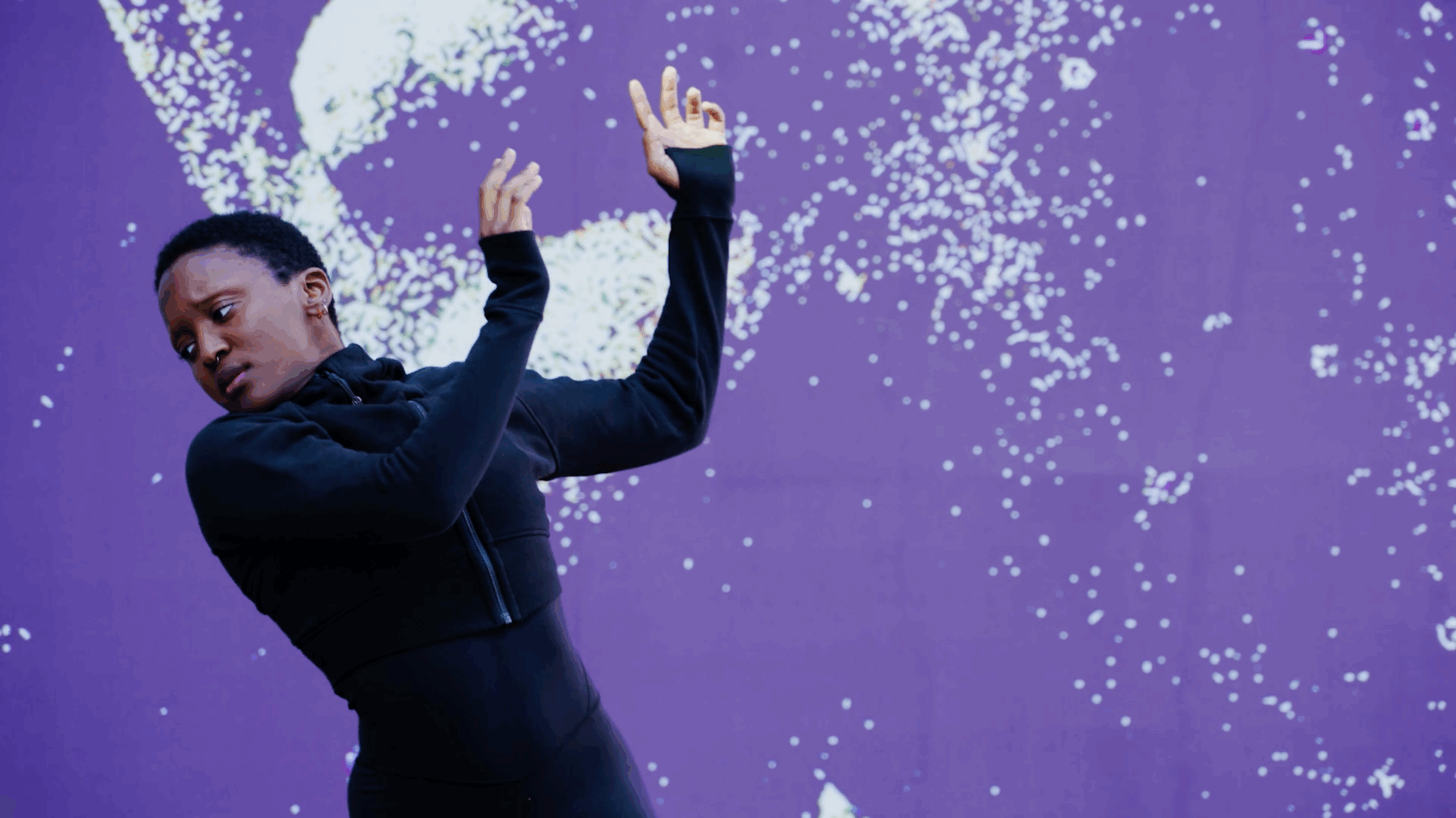 A freeze frame black woman with short hair dancing infront of a purple mural with white spots.
