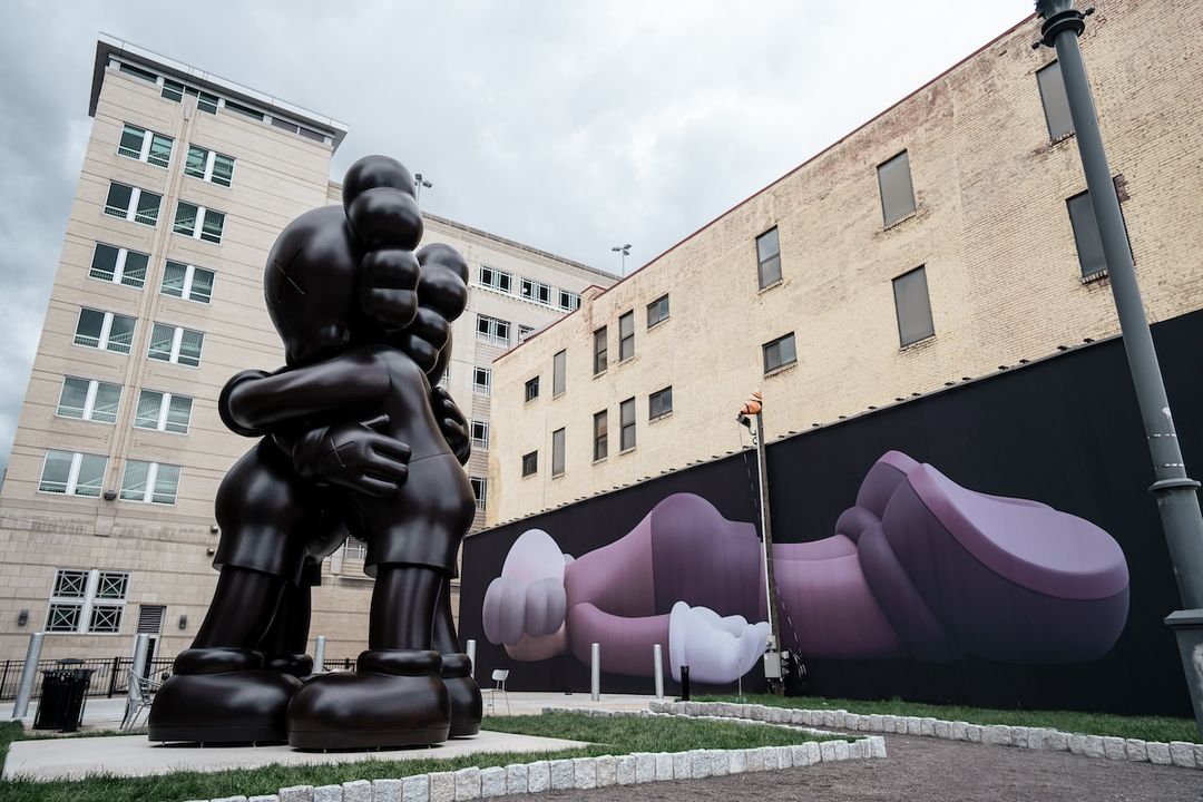 A large sculpture of two figures hugging. In the background a large mural of a figure is laying face down.