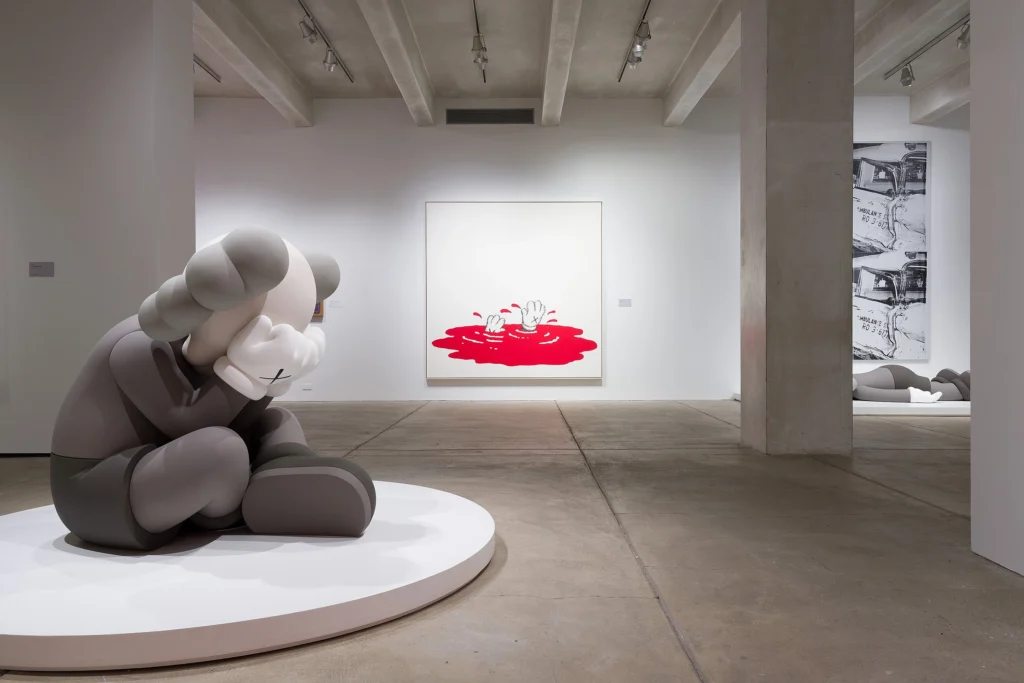 Museum installation shot of a KAWS figure sitting and the background of a cartoon pool of blood