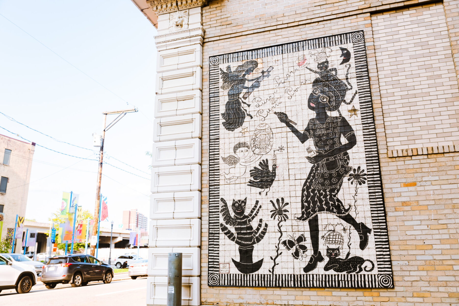 Ceramic tile mural by artist Laura Jean McLaughlin with black and white tiles depicting a woman surrounded by three to four cats.