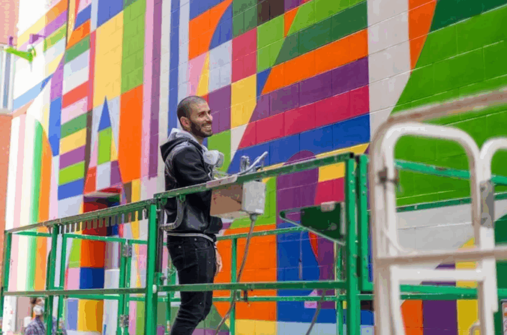 Smiling man with close shaved hair stands on scissor lift paints a rainbow colored mural