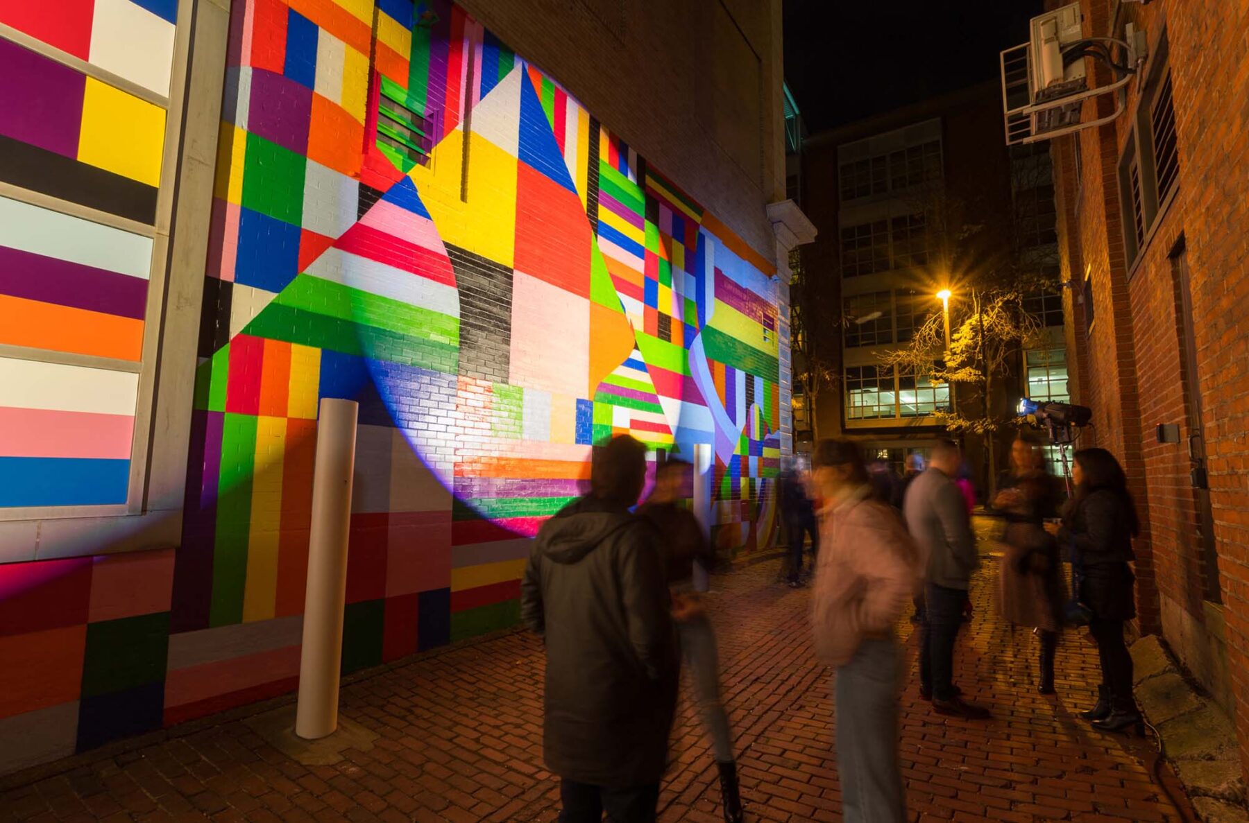 Over the Rainbow mural by Typoe in Silver Street. The image is during the night time and multiple spotlights are shining on the mural with people socializing in the foreground.