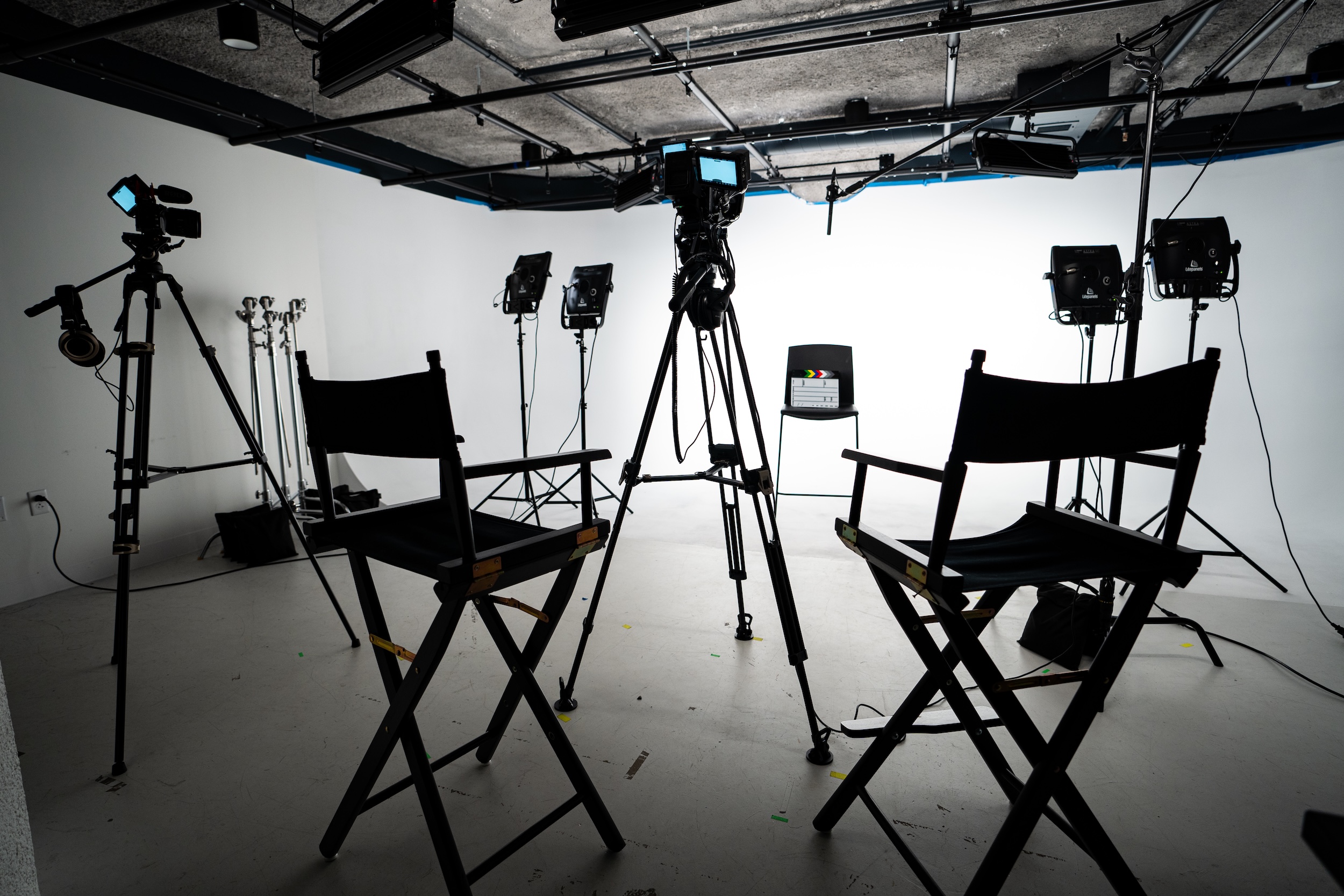An empty film studio setup featuring two director chairs facing a white backdrop, surrounded by cameras on tripods, overhead rigging, and multiple studio lights prepared for an interview or video shoot.
